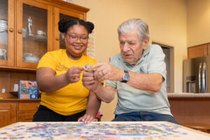 A young woman and an elderly man work together on a puzzle in a cozy kitchen, promoting dementia care and support services.