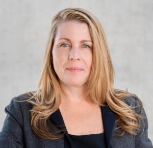 Portrait of a confident professional woman with blonde hair and blue eyes, wearing a dark blazer, against a neutral background.